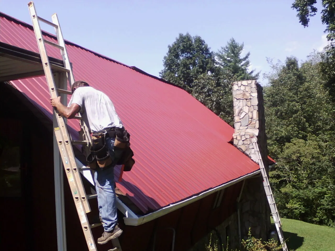Roofer on a ladder beside a red metal roof and stone chimney — Soffit & Fascia Repair by Roofing Brothers in Kensal